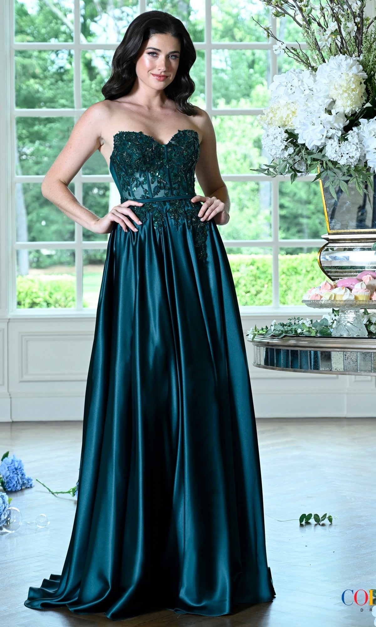 A woman poses in a deep green Long Prom Dress: Colors Dress 4025, featuring a strapless beaded bodice and a floor-length full skirt.